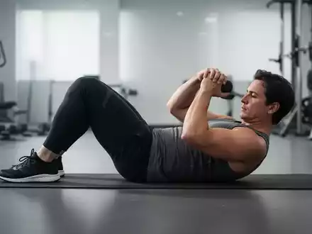 Eine Person führt auf einer Yogamatte einen modifizierten Curl-up aus, der die Rumpfmuskulatur stärkt, mit einem unscharfen Fitnessstudio im Hintergrund. - Foto: über boerse-global.de