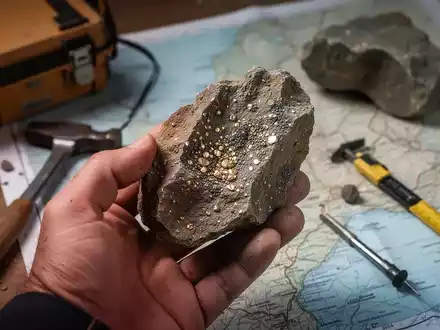 Nahaufnahme einer Hand, die einen Bohrkern mit metallischen Sprenkeln hält, im Hintergrund geologische Werkzeuge und eine Karte. - Foto: über boerse-global.de