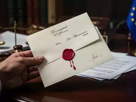 Eine Hand hält einen dringenden Brief mit rotem Wachssiegel auf einem Schreibtisch, im Hintergrund unscharf EU-Dokumente und Flagge. - Foto: über boerse-global.de