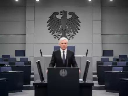 Ein unscharfer Mann steht an einem Rednerpult in einem modernen Parlamentssaal, im Hintergrund das Bundestagssymbol. - Foto: über boerse-global.de