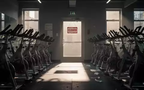 Leeres Fitnessstudio mit Trainingsgeräten, einer verschlossenen Tür und einem Schild, das auf Kursausfälle hinweist, während die Sonne durch die Fenster scheint. - Foto: über boerse-global.de