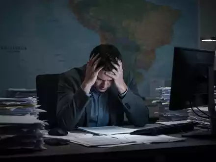Eine erschöpfte Person sitzt mit dem Kopf in den Händen an einem Schreibtisch, umgeben von Papieren und einem leuchtenden Bildschirm. - Foto: über boerse-global.de