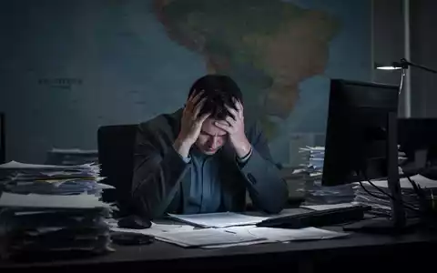Eine erschöpfte Person sitzt mit dem Kopf in den Händen an einem Schreibtisch, umgeben von Papieren und einem leuchtenden Bildschirm. - Foto: über boerse-global.de