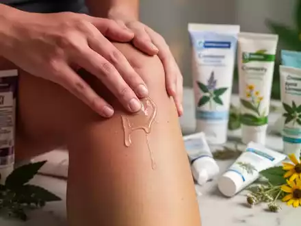 Eine Hand trägt Schmerzgel auf ein Knie auf, im Hintergrund sind verschiedene Tuben Schmerzgel und pflanzliche Inhaltsstoffe zu sehen. - Foto: über boerse-global.de