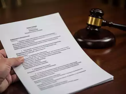 Eine Nahaufnahme einer Hand, die ein juristisches Dokument mit verschwommenem Text hält, mit einem Hammer auf einem dunklen Holztisch im Hintergrund. - Foto: über boerse-global.de