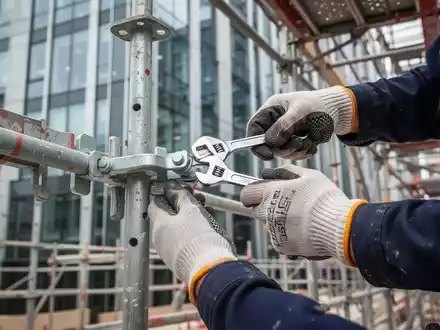 Nahaufnahme von Händen eines Bauarbeiters mit Schutzhandschuhen, der eine Gerüstklemme mit einem Schraubenschlüssel befestigt. - Foto: über boerse-global.de