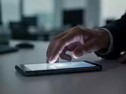 Eine Hand greift nach einem leuchtenden Smartphone auf einem Schreibtisch mit verschwommenem Bürohintergrund, was digitalen Stress symbolisiert. - Foto: über boerse-global.de