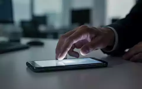 Eine Hand greift nach einem leuchtenden Smartphone auf einem Schreibtisch mit verschwommenem Bürohintergrund, was digitalen Stress symbolisiert. - Foto: über boerse-global.de