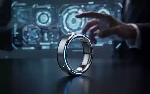 Schlanker silberner Smart-Ring mit blauen Akzenten auf dunkler Oberfläche, verschwommene Hand interagiert im Hintergrund mit holografischem Interface. - Foto: über boerse-global.de