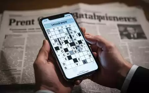 Eine Hand hält ein Smartphone mit einem digitalen Kreuzworträtsel; im Hintergrund verschwommen eine Zeitung. - Foto: über boerse-global.de