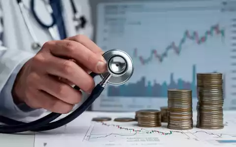 Eine Hand im Arztkittel hält ein Stethoskop, im Hintergrund verschwommene Finanzgrafiken, die finanzielle Belastung im Gesundheitswesen darstellen. - Foto: über boerse-global.de