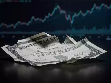 Ein zerknittertes Aktienzertifikat liegt auf einer dunklen Oberfläche, im Hintergrund verschwommene, fallende Finanzdiagramme. - Foto: über boerse-global.de