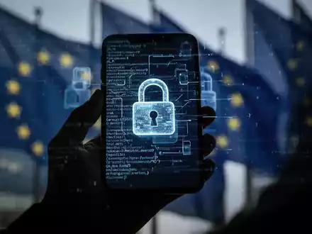 Eine Hand hält ein Smartphone mit digitalen Vorhängeschlössern und Codezeilen, im Hintergrund verschwommene EU-Flaggen. - Foto: über boerse-global.de