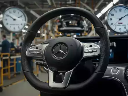 Nahaufnahme eines Mercedes-Benz Lenkrads mit verschwommenem Fabrikhintergrund und einer Uhr, die längere Arbeitszeiten anzeigt. - Foto: über boerse-global.de