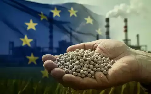 Düngemittelpellets in der Hand eines Landwirts, mit verschwommenem Hintergrund, der die EU-Flagge und Industrieschornsteine zeigt. - Foto: über boerse-global.de