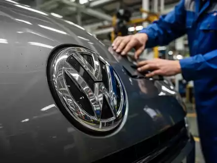 Nahaufnahme eines VW-Emblems, teilweise verdeckt von der Hand eines Arbeiters, der Sparpläne und Stellenabbau symbolisiert. - Foto: über boerse-global.de