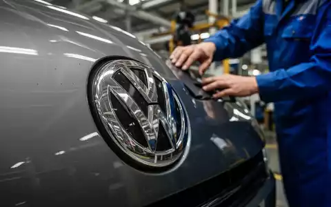 Nahaufnahme eines VW-Emblems, teilweise verdeckt von der Hand eines Arbeiters, der Sparpläne und Stellenabbau symbolisiert. - Foto: über boerse-global.de