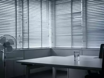 Modernes Büro mit geschlossenen Jalousien, einem Ventilator und einem Glas Wasser auf einem Schreibtisch, beleuchtet von sanftem Licht. - Foto: über boerse-global.de