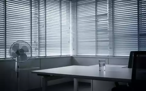 Modernes Büro mit geschlossenen Jalousien, einem Ventilator und einem Glas Wasser auf einem Schreibtisch, beleuchtet von sanftem Licht. - Foto: über boerse-global.de