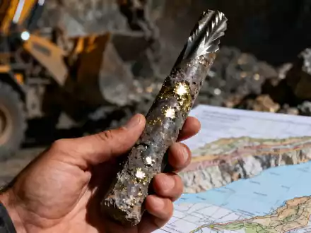 Nahaufnahme einer Hand, die eine Bohrkernprobe mit metallischen Sprenkeln hält, im Hintergrund verschwommene Bergbauausrüstung. - Foto: über boerse-global.de