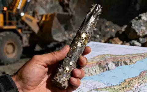 Nahaufnahme einer Hand, die eine Bohrkernprobe mit metallischen Sprenkeln hält, im Hintergrund verschwommene Bergbauausrüstung. - Foto: über boerse-global.de