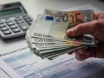 Eine Nahaufnahme einer Hand, die Euro-Banknoten hält, mit einem unscharfen Taschenrechner und einem Rentenbescheid im Hintergrund. - Foto: über boerse-global.de