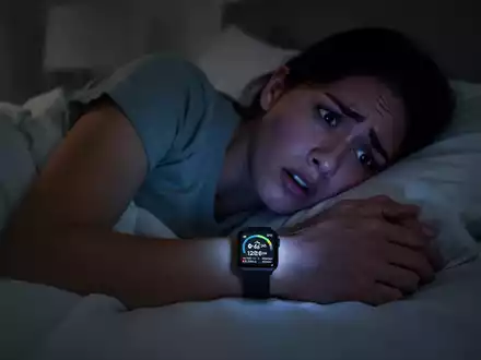 Eine Person liegt wach und ängstlich im Bett, beleuchtet vom Display einer Smartwatch, die Schlafdaten anzeigt. - Foto: über boerse-global.de