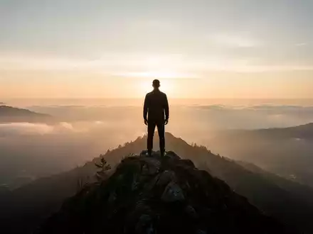 Eine Silhouette steht auf einem Berggipfel bei Sonnenaufgang und blickt auf eine weite, neblige Landschaft, die mentale Resilienz symbolisiert. - Foto: über boerse-global.de