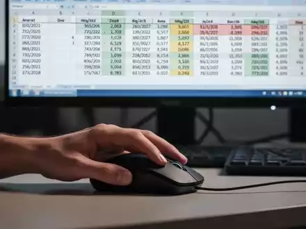 Eine Hand bedient eine Computermaus, im Hintergrund ein Monitor mit einem Tabellenkalkulationsprogramm, das verschiedene Datumsformate und Daten anzeigt. - Foto: über boerse-global.de