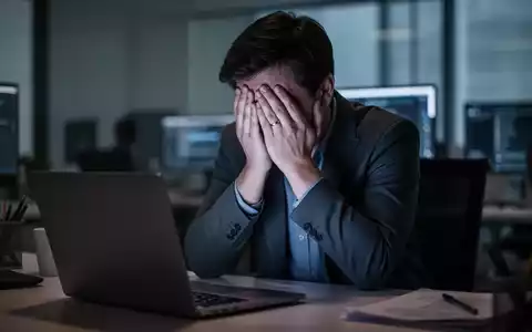 Eine Person vergräbt ihr Gesicht in den Händen, beleuchtet vom Laptop-Bildschirm in einem Büro, was digitalen Stress symbolisiert. - Foto: über boerse-global.de