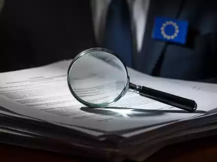 Ein Vergrößerungsglas liegt auf einem Stapel juristischer Dokumente, im Hintergrund ein Anzug mit EU-Anstecker, Fokus auf rechtliche Prüfung. - Foto: über boerse-global.de