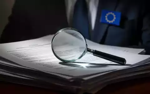 Ein Vergrößerungsglas liegt auf einem Stapel juristischer Dokumente, im Hintergrund ein Anzug mit EU-Anstecker, Fokus auf rechtliche Prüfung. - Foto: über boerse-global.de