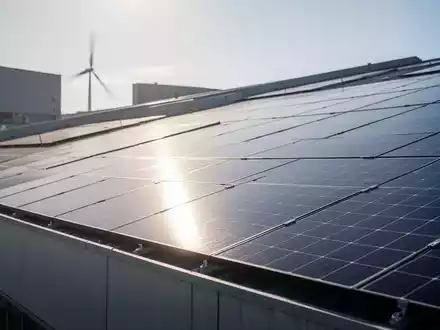 Solarpanels auf dem Dach eines Industriegebäudes, im Hintergrund schemenhaft eine Windkraftanlage. Symbolisiert erneuerbare Energien für Unternehmen. - Foto: über boerse-global.de