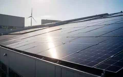 Solarpanels auf dem Dach eines Industriegebäudes, im Hintergrund schemenhaft eine Windkraftanlage. Symbolisiert erneuerbare Energien für Unternehmen. - Foto: über boerse-global.de