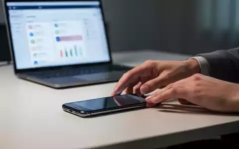 Eine Person legt ein Smartphone bewusst verdeckt auf einen Schreibtisch, mit einem unscharfen Laptop im Hintergrund. - Foto: über boerse-global.de