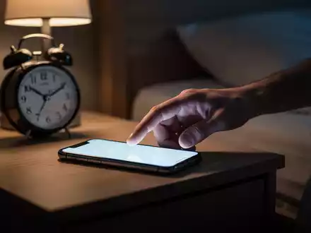 Eine Hand greift nach einem Smartphone auf einem Nachttisch in einem abgedunkelten Schlafzimmer, das digitale Erschöpfung symbolisiert. - Foto: über boerse-global.de