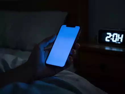 Eine Hand hält ein leuchtendes Smartphone in einem dunklen Schlafzimmer; die Uhrzeit auf dem Bildschirm ist spät in der Nacht. - Foto: über boerse-global.de