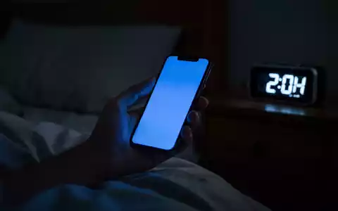 Eine Hand hält ein leuchtendes Smartphone in einem dunklen Schlafzimmer; die Uhrzeit auf dem Bildschirm ist spät in der Nacht. - Foto: über boerse-global.de