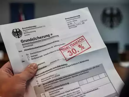 Eine Nahaufnahme einer Hand, die einen deutschen Grundsicherungsbescheid mit einem roten Sanktionsstempel hält. - Foto: über boerse-global.de