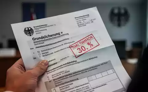 Eine Nahaufnahme einer Hand, die einen deutschen Grundsicherungsbescheid mit einem roten Sanktionsstempel hält. - Foto: über boerse-global.de
