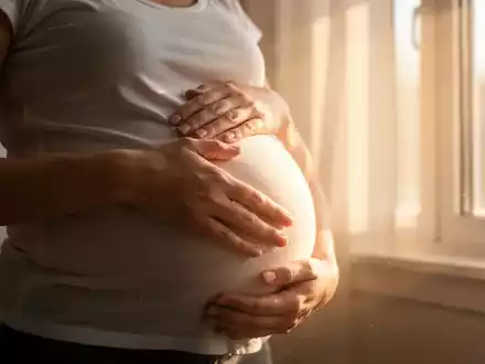 Eine schwangere Frau berührt ihren Bauch, mit Sonnenlicht, das durch ein Fenster fällt und sie warm beleuchtet. - Foto: über boerse-global.de