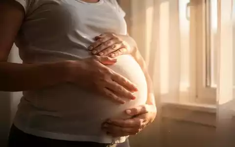 Eine schwangere Frau berührt ihren Bauch, mit Sonnenlicht, das durch ein Fenster fällt und sie warm beleuchtet. - Foto: über boerse-global.de