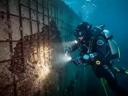 Ein Industrietaucher inspiziert unter Wasser eine beschädigte Kaimauer mit Rissen und Korrosion, beleuchtet von einem hellen Scheinwerfer. - Foto: über boerse-global.de