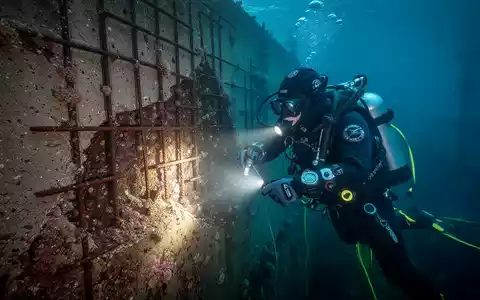 Ein Industrietaucher inspiziert unter Wasser eine beschädigte Kaimauer mit Rissen und Korrosion, beleuchtet von einem hellen Scheinwerfer. - Foto: über boerse-global.de