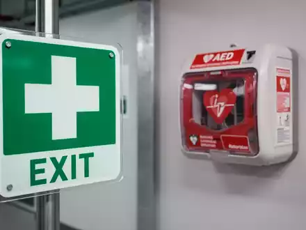 Ein deutlich sichtbares Sicherheitszeichen im Vordergrund und ein Defibrillator im unscharfen Hintergrund in einer modernen Umgebung. - Foto: über boerse-global.de