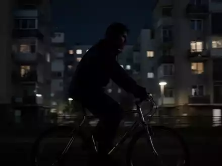 Eine verschwommene, dunkle Figur fährt nachts auf einem Fahrrad durch eine Stadt, im Hintergrund verschwommene Gebäude. - Foto: über boerse-global.de