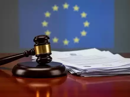 Ein stilisierter Rechtshammer auf einem Holztisch, umgeben von Dokumenten, mit einer angedeuteten EU-Flagge im Hintergrund. - Foto: über boerse-global.de