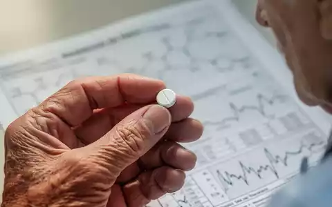Eine Nahaufnahme der Hand einer älteren Person, die eine weiße Pille hält, mit einem unscharfen medizinischen Diagramm im Hintergrund. - Foto: über boerse-global.de