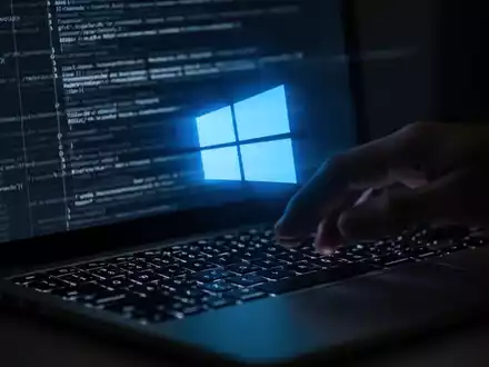 Eine Hand schwebt über einer Laptop-Tastatur, auf dem Bildschirm leuchtet ein blaues Windows-Logo und abstrakter Code. - Foto: über boerse-global.de