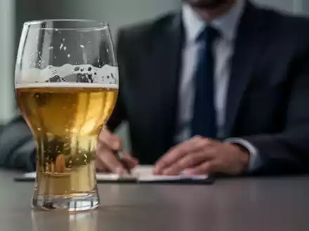 Ein unscharfes Glas Bier oder Wein auf einem Tisch, im Hintergrund eine Person in formeller Kleidung, die ein Vorstellungsgespräch andeutet. - Foto: über boerse-global.de